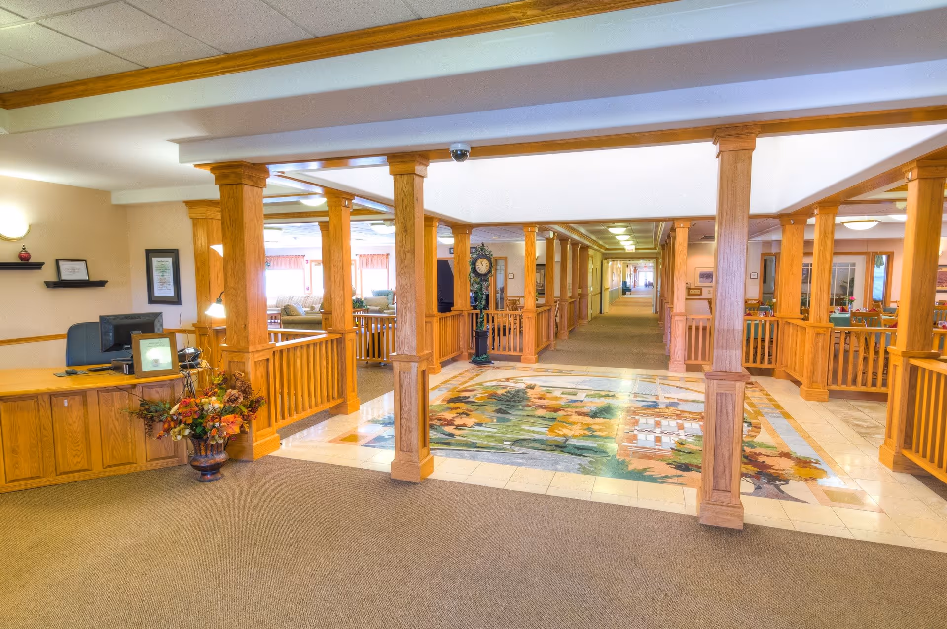 Interior view of a senior living facility lobby area with wooden pillars and railings, a colorful tiled floor section with a nature-themed design, a reception desk with a computer and framed certificates, and a long hallway extending into the distance. There are seating areas with couches and a grandfather clock visible in the background.