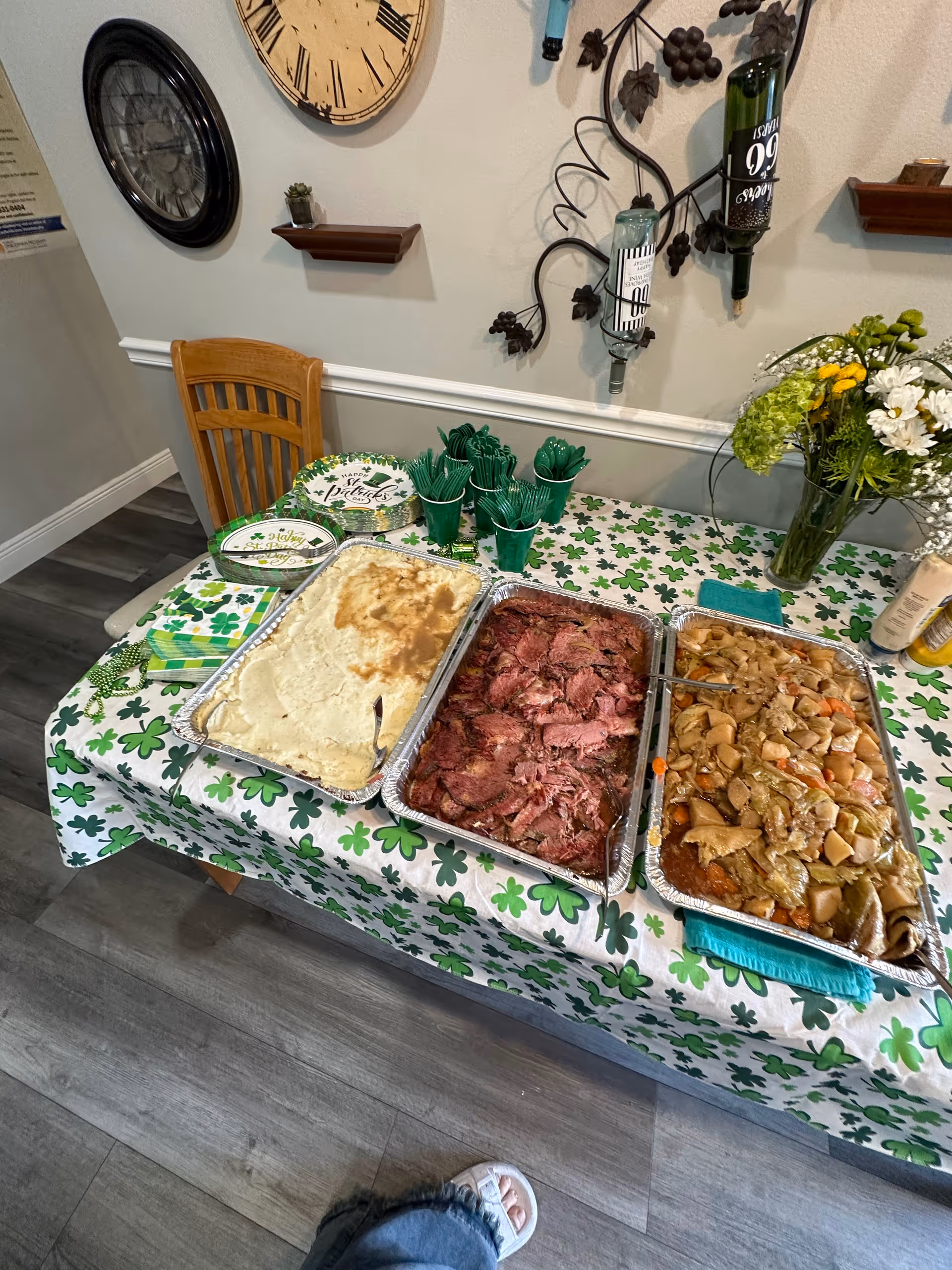 A dining table set with trays of mashed potatoes, sliced roast beef and stewed vegetables on a shamrock tablecloth with green utensils and floral decor.
