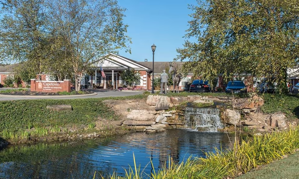 Exterior view of Capetown Senior Living facility with a pond and small waterfall in the foreground, trees on both sides, a brick sign with the facility name, and a building entrance with an American flag and parked cars.
