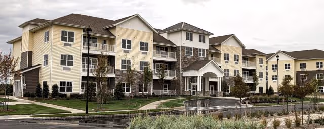 Exterior view of a large, multi-story senior living facility named The Chelsea at Brookhaven, featuring beige and brown siding, multiple balconies, a covered entrance, landscaped grounds, and a circular driveway.