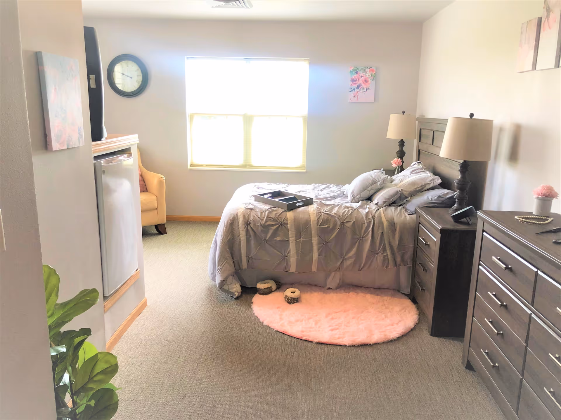 A bright bedroom with a bed covered in gray bedding and multiple pillows, a pink round rug on the floor, a wooden nightstand with a lamp and decorative items, a matching dresser with drawers, a yellow armchair near a window, a wall clock, and some floral wall art.