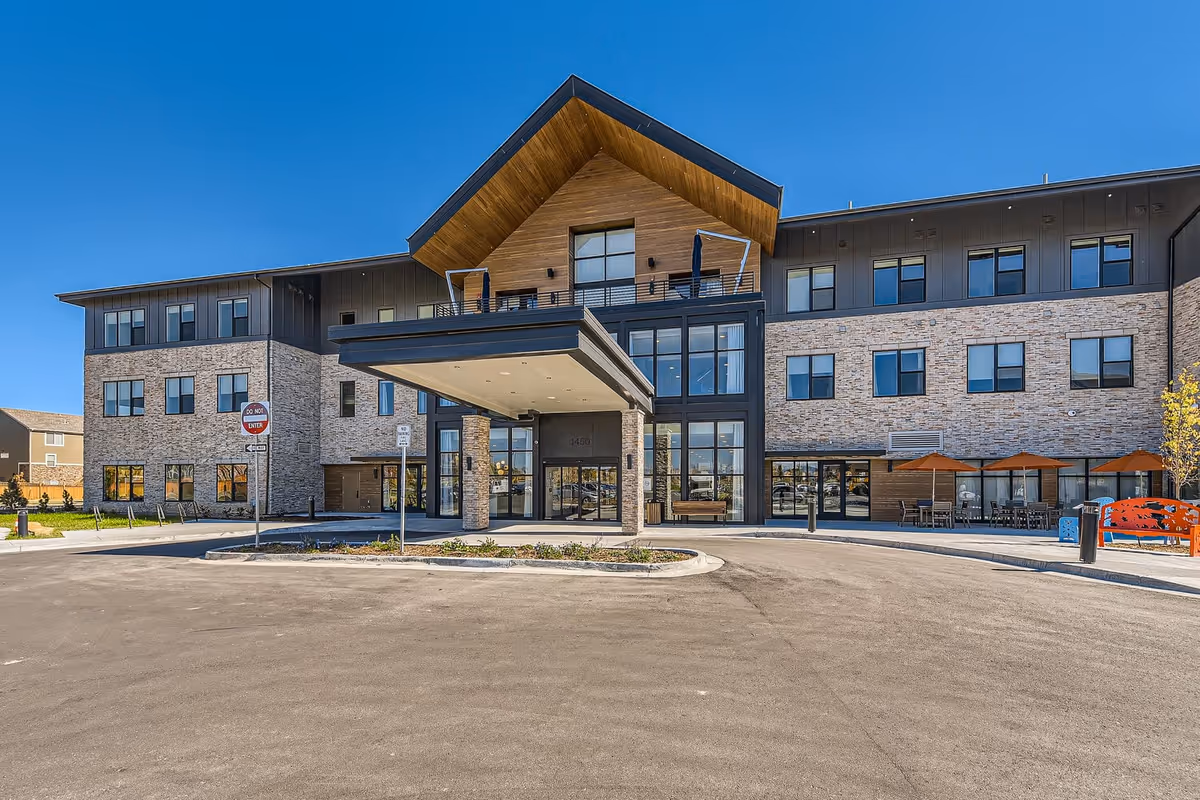 Front exterior view of The Gallery at Broomfield Assisted & Senior Living building with a covered entrance, large windows, and outdoor seating area with orange umbrellas under a clear blue sky.