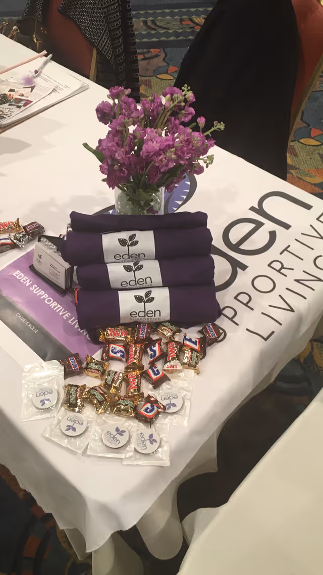 Table display with purple flowers, folded Eden-branded cloths and candy on an Eden Supportive Living tablecloth.