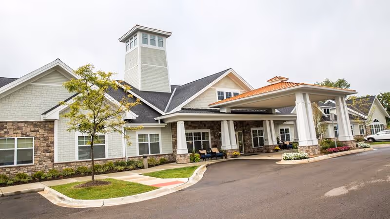 Exterior view of Pomeroy Senior Living of Northville building showing a covered entrance with white pillars, stone and light green siding, a small tree, and a driveway under an overcast sky.