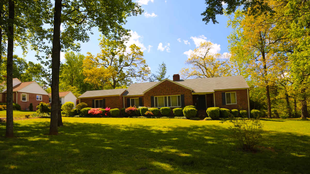 A single-story brick house with a gray roof surrounded by green grass, bushes, and trees under a blue sky with some clouds.