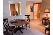 Interior view of a senior living facility dining area with a small round wooden table set for two, two wooden chairs, a side table with a lamp, and a hallway leading to another room with floral curtains.