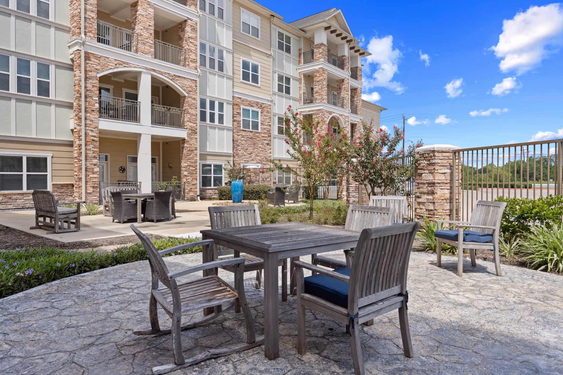 Outdoor patio area at Discovery Village Clear Lake with wooden tables and chairs on a stone-paved surface, surrounded by greenery and flowering bushes. The multi-story building with stone and beige siding is visible in the background under a blue sky with scattered clouds.