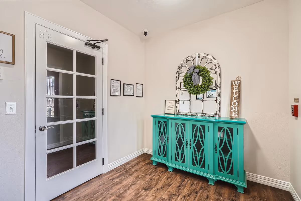 Interior corner of a senior living facility with a white door featuring glass panels on the left, a turquoise cabinet with decorative cutouts on the right, a rustic mirror with a green wreath hanging above the cabinet, and a vertical wooden sign that says 'WELCOME'. The floor is wooden, and the walls are light-colored with some framed certificates and a security camera in the corner.