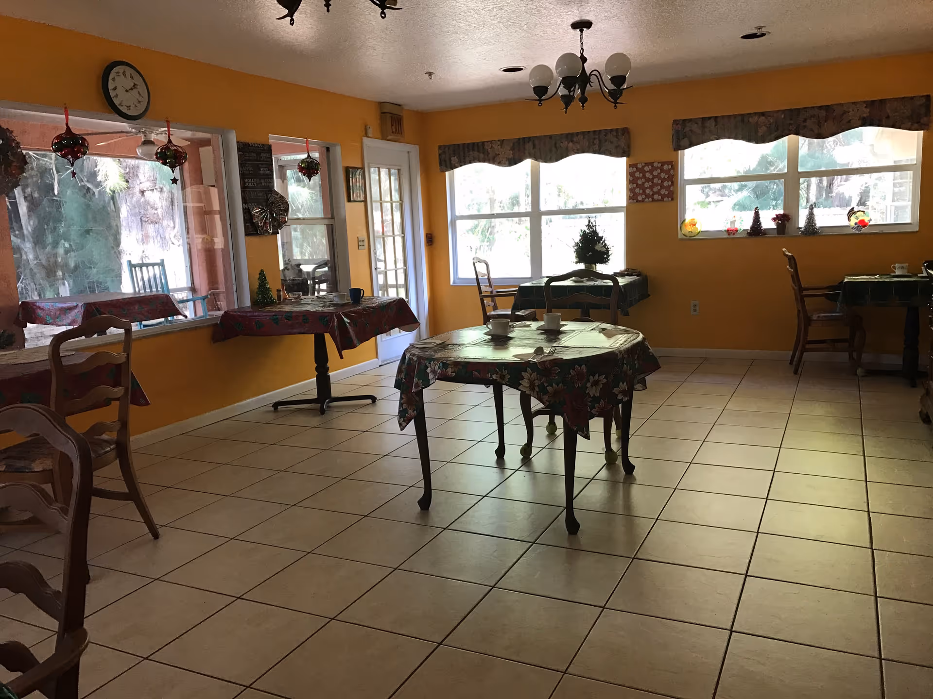 A dining room with yellow walls and tiled floor featuring several tables covered with floral tablecloths. Each table has chairs around it and some have cups and small decorative items. Large windows allow natural light to enter, and there are hanging light fixtures on the ceiling. A clock is mounted on the wall near a door leading outside.