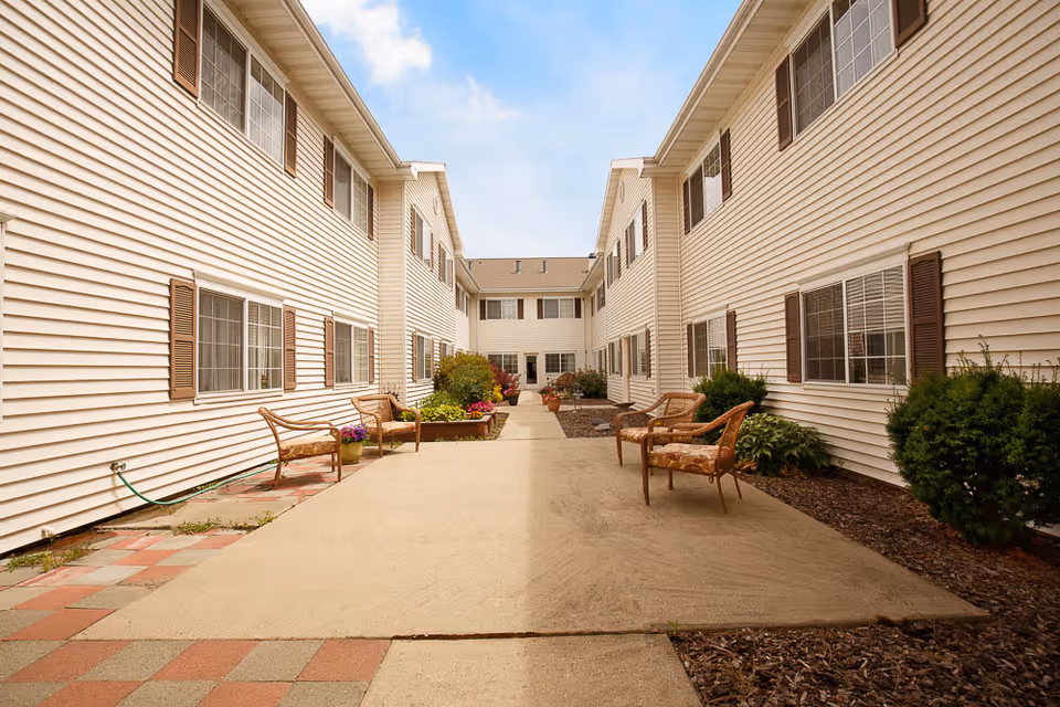 Outdoor courtyard area between two beige two-story buildings with multiple windows and brown shutters. The courtyard has a paved walkway with several cushioned chairs and benches, along with some potted plants and shrubs. The sky is partly cloudy.