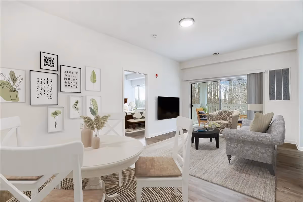 A bright and modern living and dining area with white walls and wooden flooring. The dining area features a round white table with four white chairs, two vases with dried flowers on the table, and framed botanical and alphabet artwork on the wall. The living area has a gray patterned sofa, a round black coffee table with books and a small plant, and a patterned armchair near large sliding glass doors that open to a balcony with outdoor seating and a view of trees. A flat-screen TV is mounted on the wall between the two areas.