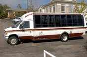 A white-and-maroon shuttle bus parked in front of a multi-story senior living building with a covered entrance.