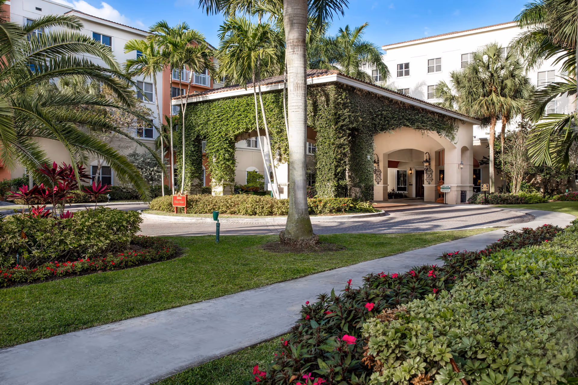 Exterior view of The Palace Renaissance & Royale senior living facility showing a covered entrance with ivy-covered arches, surrounded by palm trees, manicured lawns, and colorful flower beds under a blue sky.