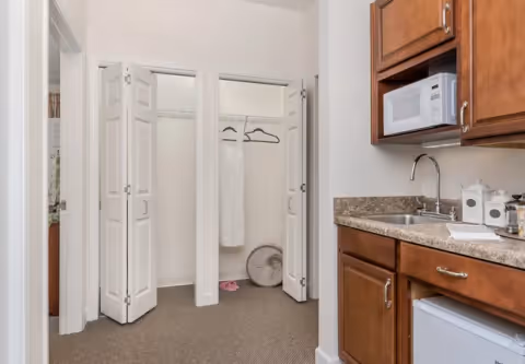 Interior view of a room with two open closets showing hanging clothes and a fan on the floor. To the right, there is a kitchenette area with a sink, countertop, microwave, and wooden cabinets.