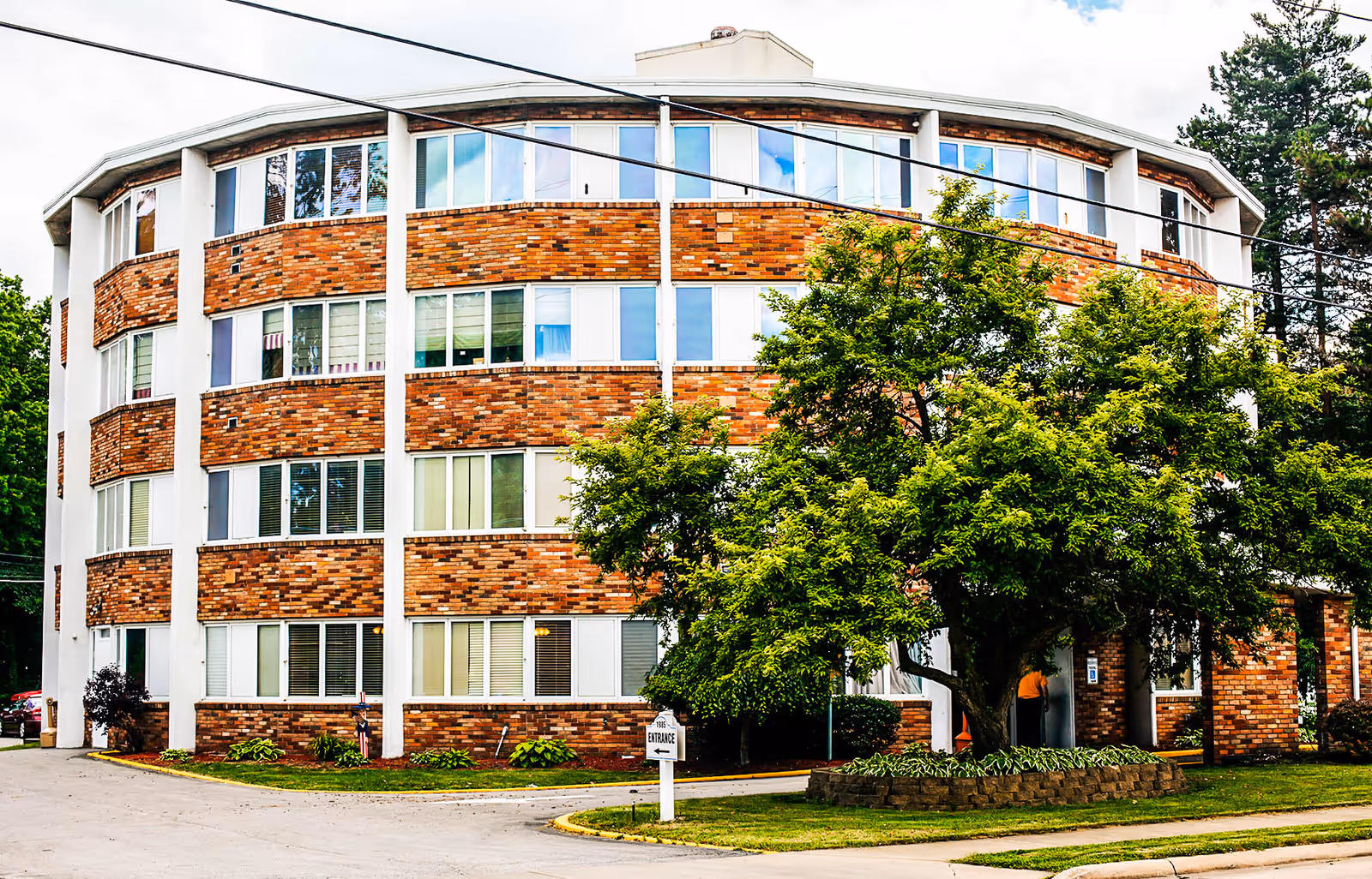 Exterior view of a multi-story senior living facility building with a brick and white facade, surrounded by trees and greenery. A driveway and a sign indicating the entrance are visible in front of the building.