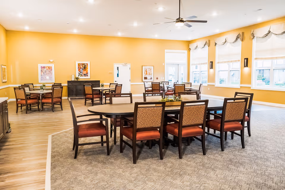 A spacious dining room with multiple tables and chairs arranged neatly. The walls are painted yellow and decorated with framed artwork. Large windows with white blinds allow natural light to fill the room. A ceiling fan with a light fixture hangs from the ceiling.