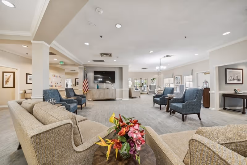 A spacious and well-lit senior living facility common area with beige and blue upholstered chairs and sofas arranged around small tables. A flat-screen TV is mounted on the wall above a cabinet, and an American flag stands nearby. The room features neutral-colored walls, carpeted floors, framed artwork, and large windows allowing natural light to enter. A floral arrangement is placed on a round table in the foreground.