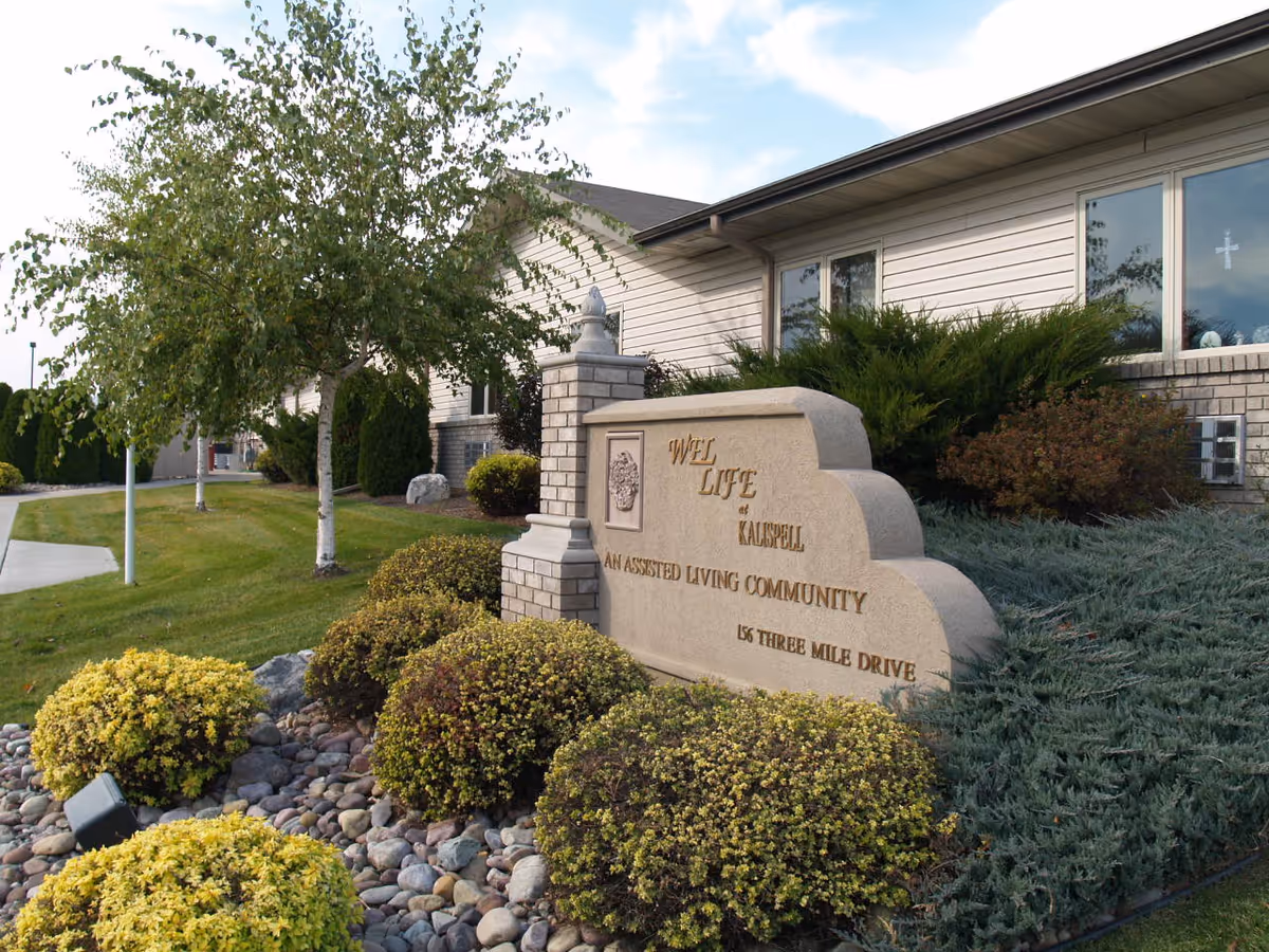 Exterior view of WEL-Life at Kalispell assisted living community showing a stone sign with the facility name and address surrounded by landscaped bushes and trees, with part of the building visible in the background under a partly cloudy sky.