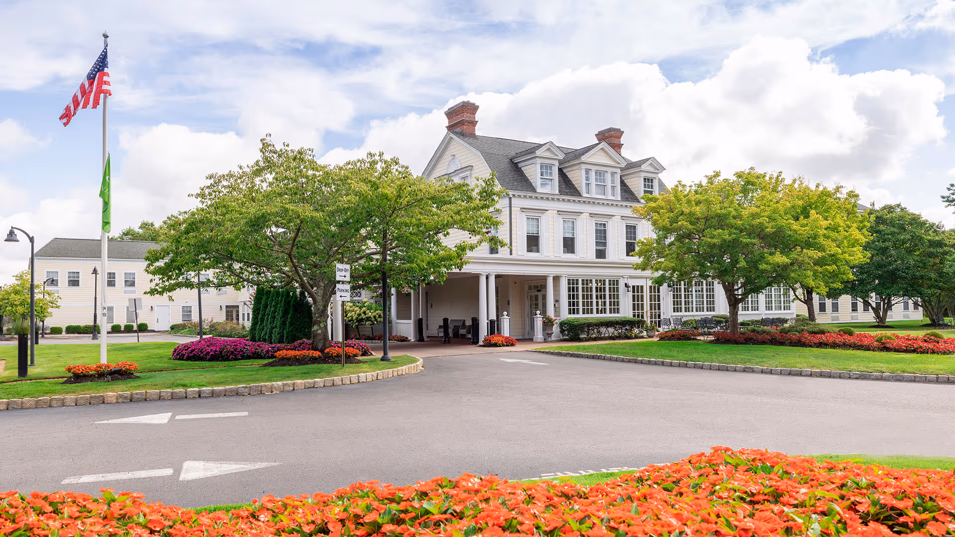 Exterior view of Atria Bay Shore facility showing a large white multi-story building with a covered entrance, surrounded by well-maintained green lawns, trees, and colorful flower beds. An American flag and a green flag are flying on flagpoles near the driveway.