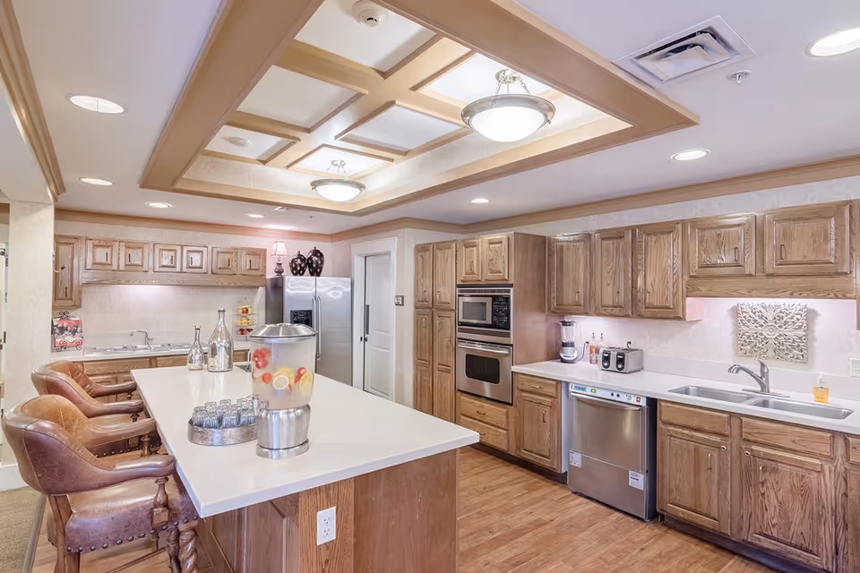 Well-lit communal kitchen with a large island and barstools, wooden cabinets, and stainless steel appliances.