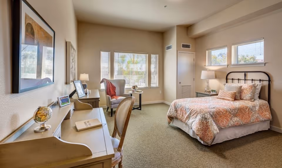 A bright and cozy bedroom with a bed featuring an orange and white patterned comforter, a bedside table with a lamp, a desk with a chair and decorative items including a small globe, and a comfortable armchair near windows that let in natural light.