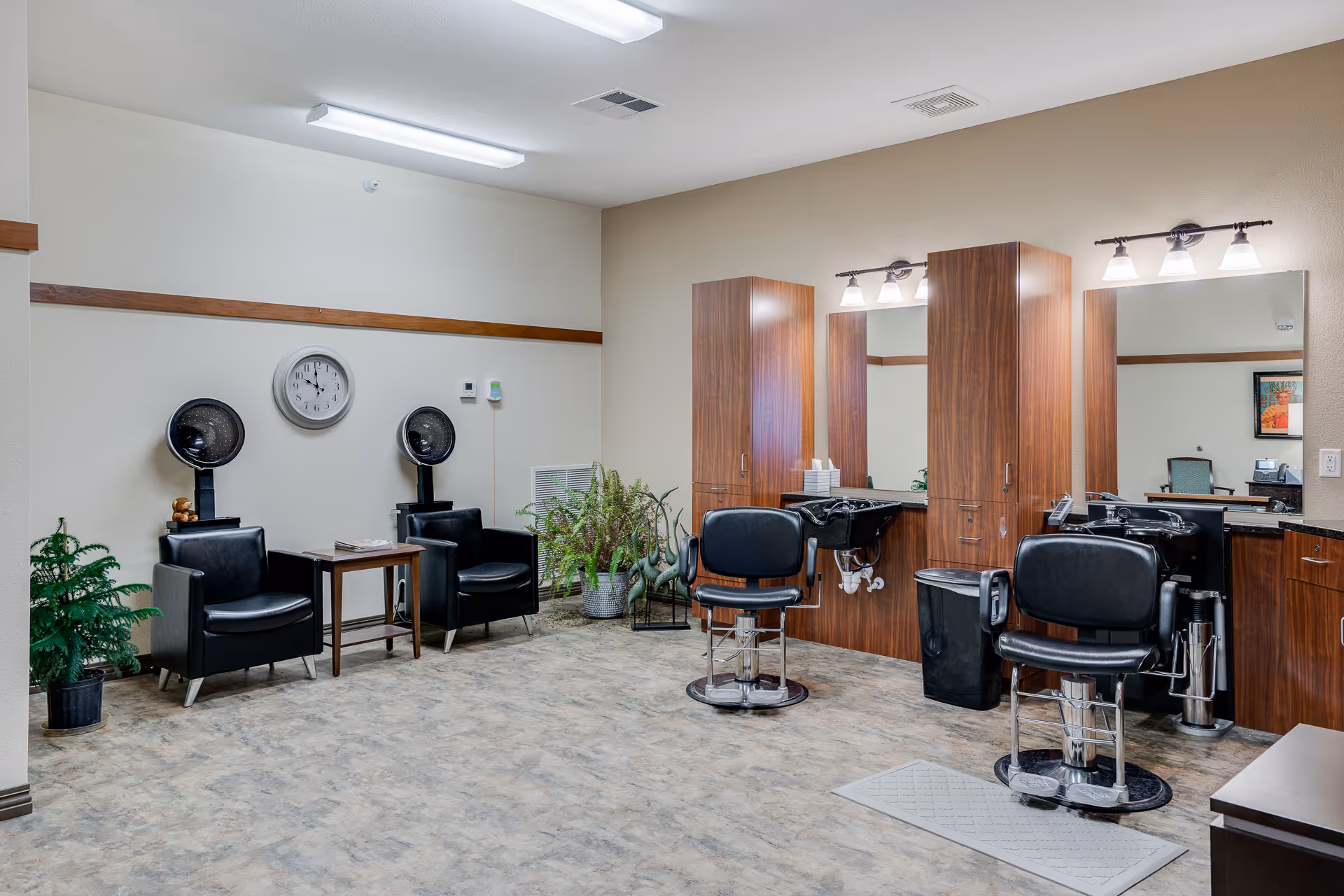 Interior of a senior living facility hair salon with two black salon chairs in front of large mirrors and wooden cabinets. Two black hair dryer chairs are positioned against the opposite wall under a clock. There are several potted plants and a small table between the hair dryer chairs. The room has bright overhead lighting and a clean, spacious floor.