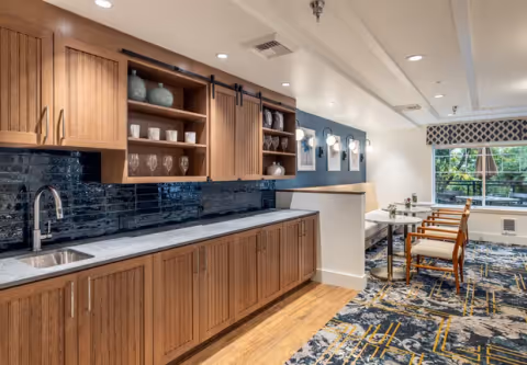 Interior view of a senior living facility kitchen area with wooden cabinets, a marble countertop, a sink, and glassware displayed on open shelves. Adjacent to the kitchen area is a dining space with tables and chairs, a large window with a patterned valance, and wall-mounted lights.