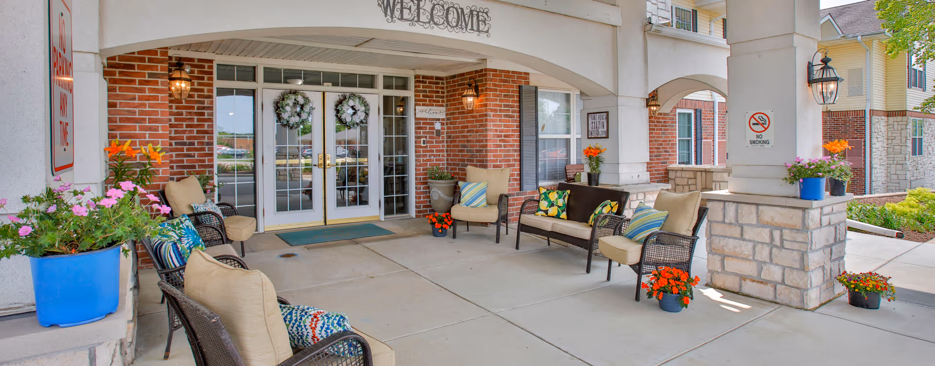 Covered entrance area of a senior living facility with cushioned wicker chairs and a loveseat arranged around the space. The entrance features double glass doors decorated with wreaths, brick walls, stone pillars, and various potted flowers. Signs indicating no parking and no smoking are visible on the pillars.