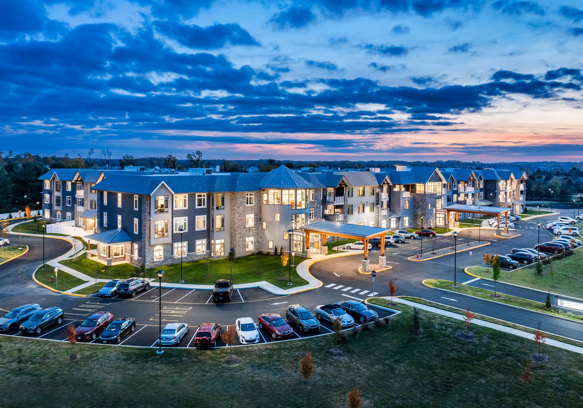 Aerial view of Meadowcrest at Middletown senior living facility at dusk, showing a large, modern three-story building with multiple windows lit from inside. The building has a mix of stone and siding exterior with peaked roofs and covered entrances. There are several cars parked in the parking lot surrounding the building, and the sky is partly cloudy with a colorful sunset in the background.