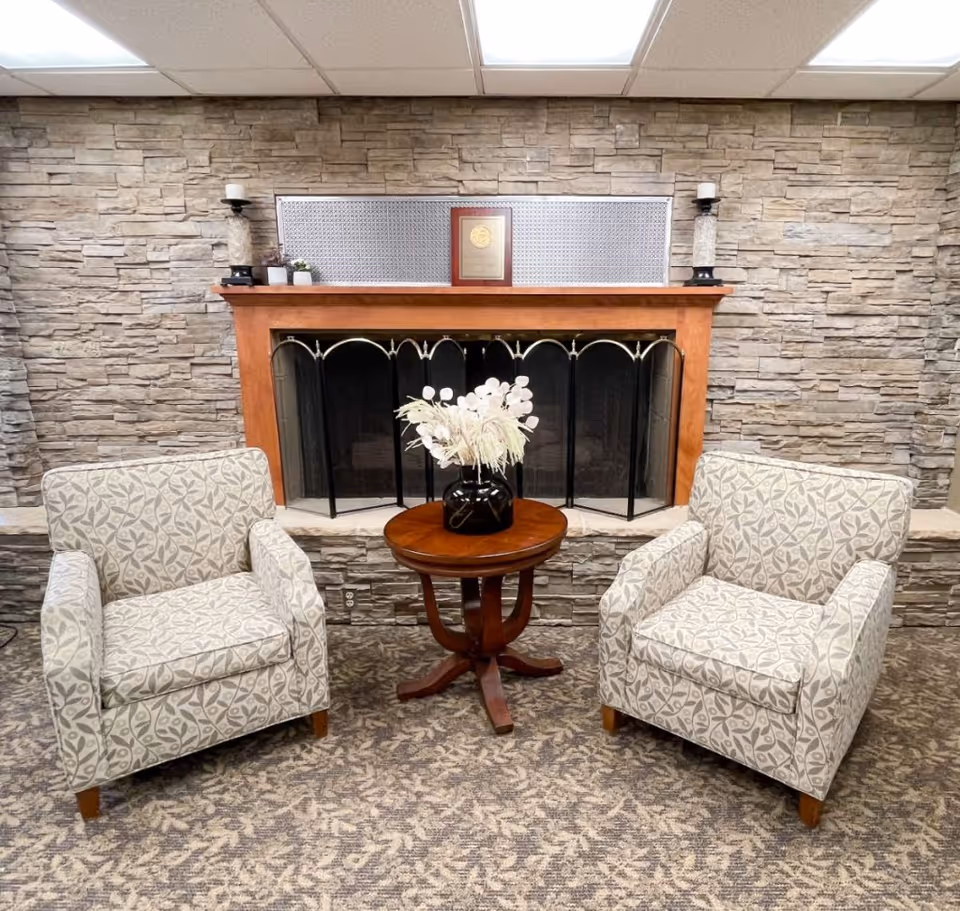 Two patterned armchairs facing each other with a small round wooden table in between, holding a black vase with white decorative flowers. Behind them is a stone wall with a wooden fireplace mantel, a fireplace screen, and decorative items including candles and a plaque on the mantel.
