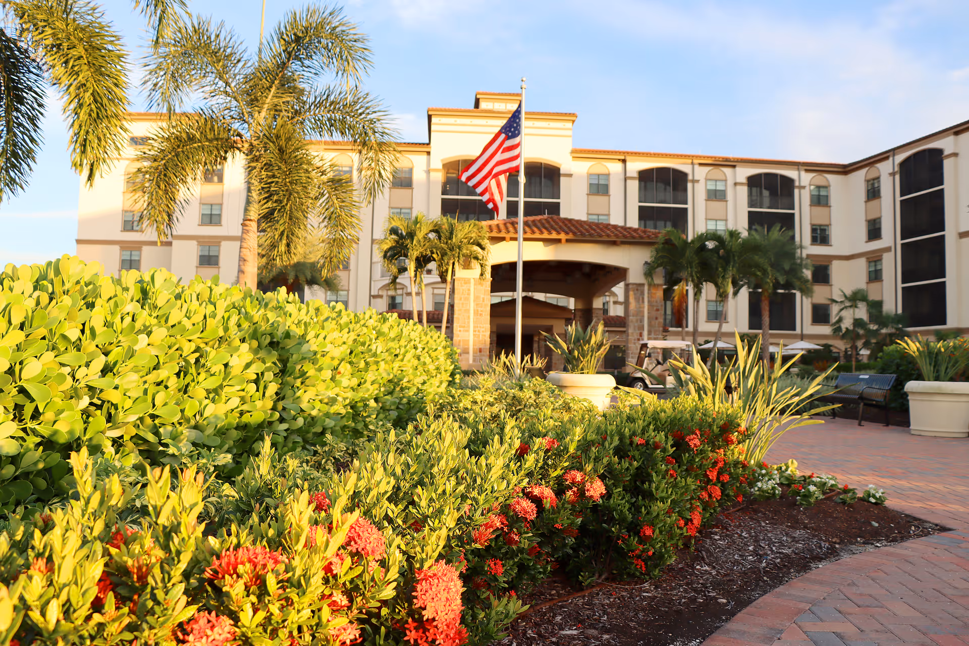 Front entrance of a multi-story residential building with palm trees, manicured shrubs, red flowers, and an American flag.