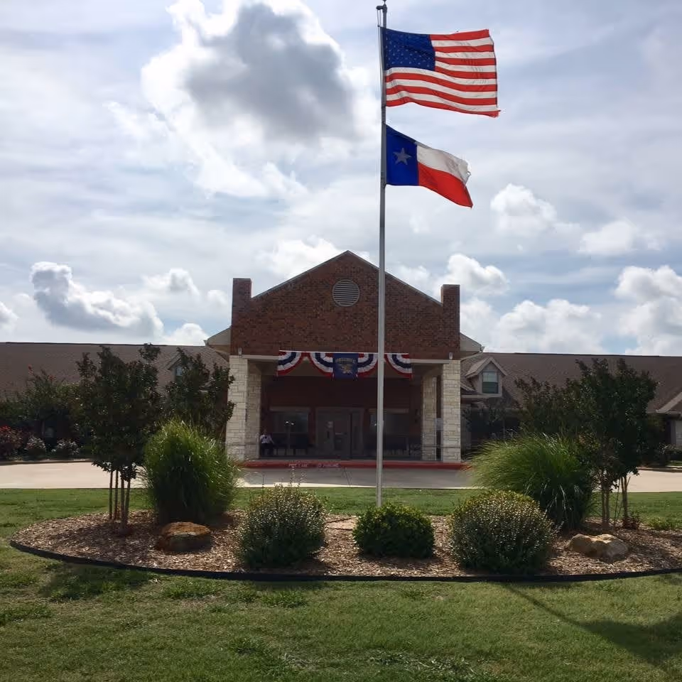 Front exterior view of Legend Oaks Healthcare and Rehabilitation - Ennis building with an American flag and Texas state flag flying on a flagpole in front. The building has a brick facade with a covered entrance decorated with red, white, and blue bunting. There are shrubs and landscaping in front of the building under a partly cloudy sky.