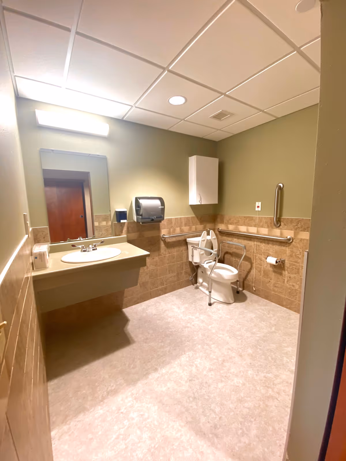 A clean and accessible bathroom with beige tiled walls and light green paint above. The room features a wall-mounted sink with a mirror and light above it on the left side. On the right side, there is a toilet equipped with safety rails and a raised toilet seat. A paper towel dispenser and a white wall cabinet are mounted on the wall above the toilet. The floor is covered with light-colored vinyl flooring.