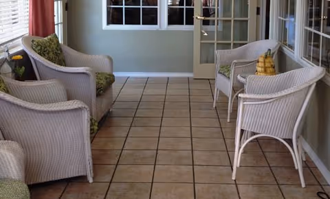 A sunlit indoor seating area with four white wicker chairs arranged along the walls, two on each side. The floor is tiled with square beige tiles, and there is a window with white blinds on the left side. A small table with a decorative item is placed near the chairs on the right side. The walls are painted light green, and there is a glass door at the end of the room.