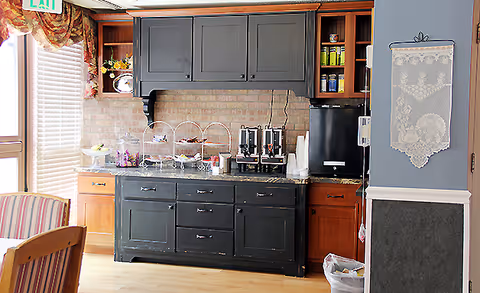 A kitchenette area with black and wooden cabinets, a granite countertop holding coffee dispensers, cups, and a three-tiered tray with snacks. To the left, there is a window with blinds and floral curtains, and a striped chair is partially visible. On the right wall, a decorative lace wall hanging is displayed.