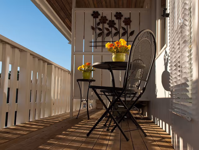 A sunlit wooden balcony with a small black metal table and two matching chairs. On the table and a small side table are green pots with bright yellow and orange flowers. The balcony has white wooden railings and a decorative panel with flower designs on the far wall. Window blinds cast shadows on the right side wall.