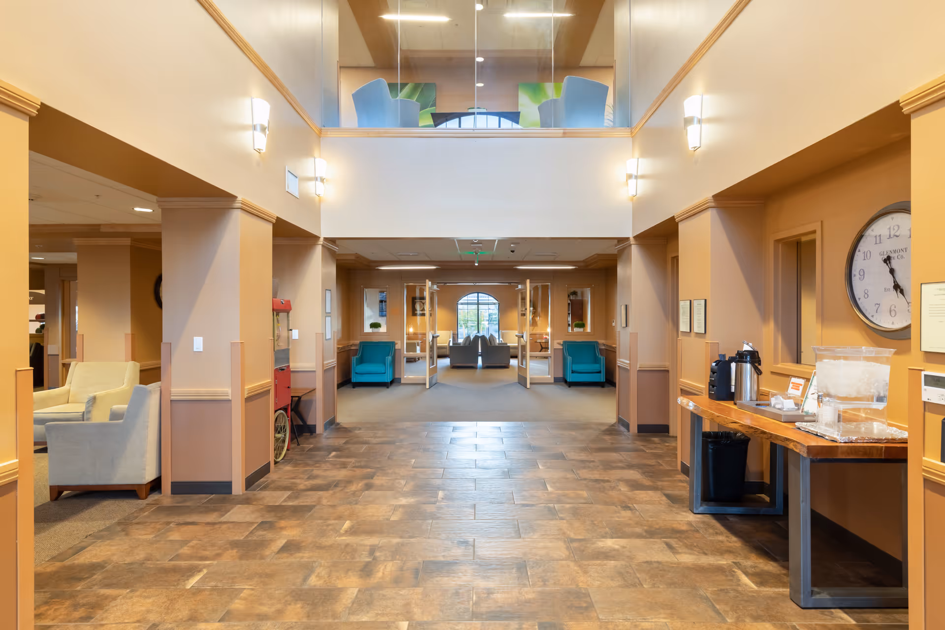 Spacious, warmly lit senior living lobby with seating areas, a tiled floor, and a refreshment table along the right wall.