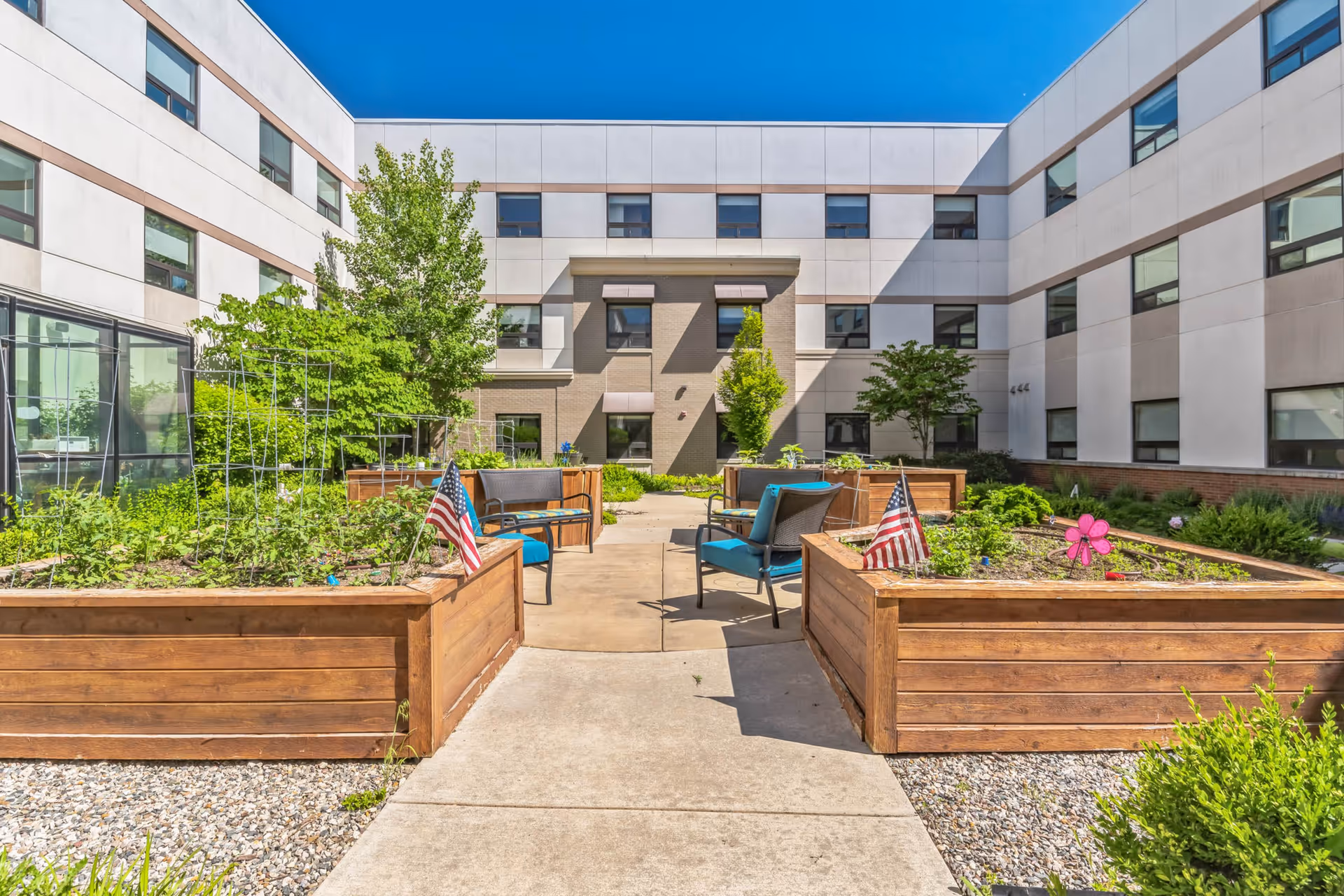 Outdoor courtyard garden area at Willowbrook Hills with raised wooden garden beds containing plants and small American flags. There are blue cushioned chairs and benches arranged around the garden beds. The courtyard is surrounded by a three-story building under a clear blue sky.