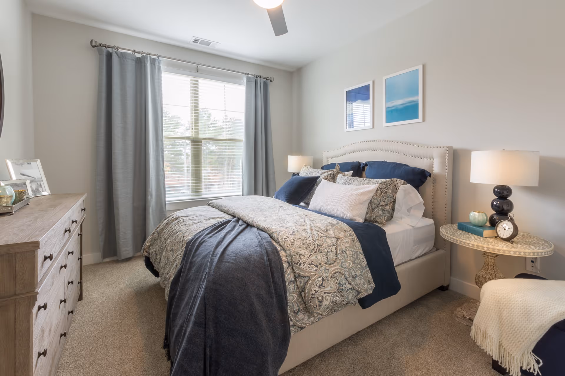 A cozy bedroom with a large bed featuring a beige upholstered headboard, multiple pillows, and a patterned comforter in shades of blue and beige. There is a wooden dresser with framed photos on the left side and a round side table with a lamp, clock, and decorative items on the right. A window with gray curtains allows natural light to fill the room.