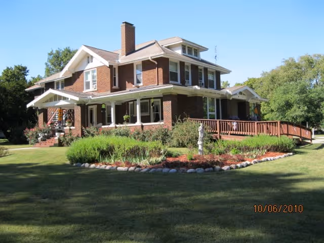 A large two-story brick house with a wraparound porch and a wheelchair ramp, surrounded by a well-maintained lawn and garden with shrubs and a small statue, under a clear blue sky.