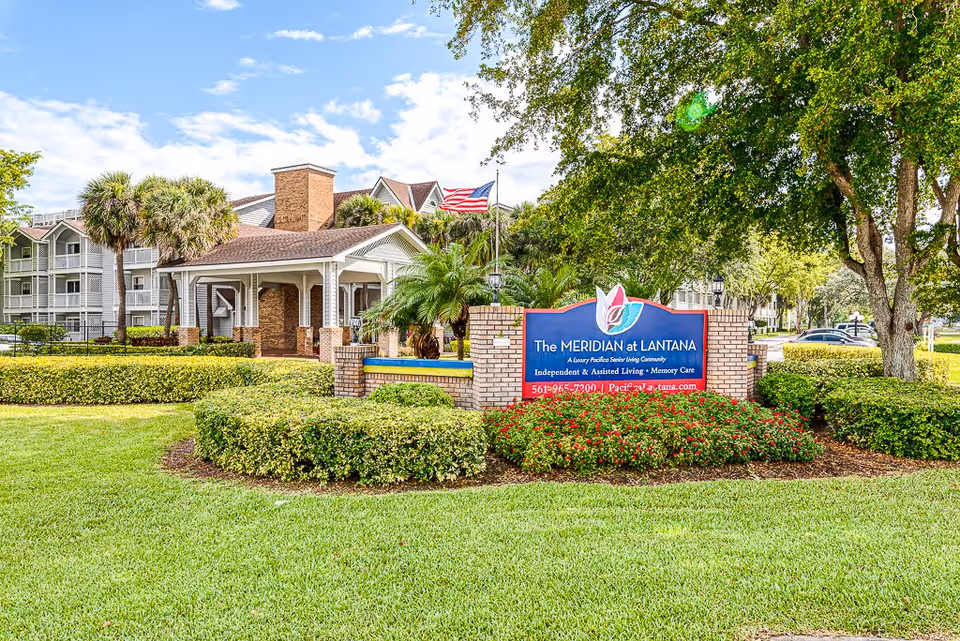 Exterior view of The Meridian at Lantana senior living community featuring a large sign with the community name, surrounded by well-maintained landscaping including bushes, flowers, and trees. The building and an American flag are visible in the background under a partly cloudy sky.