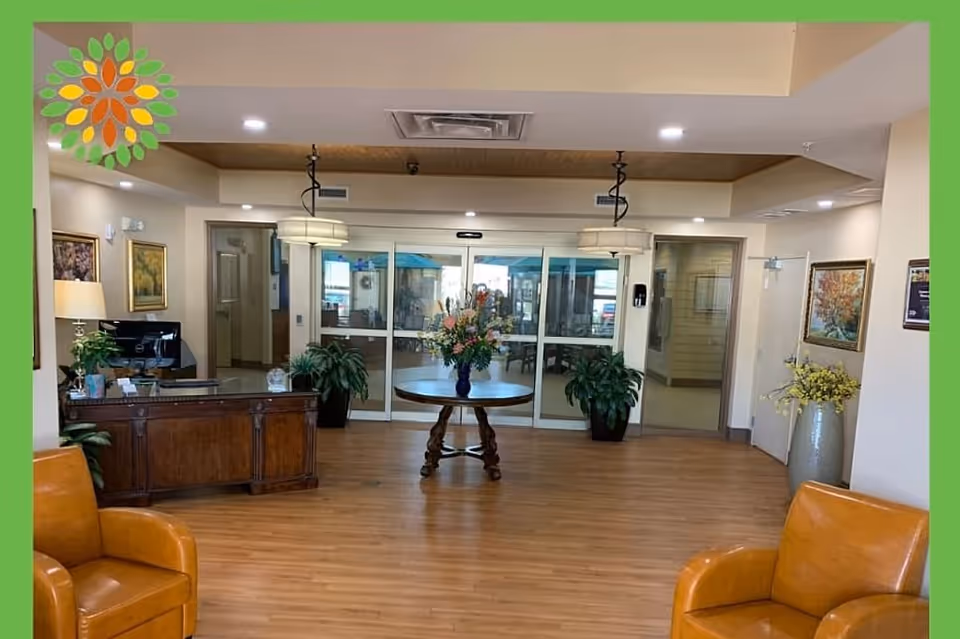 A spacious and well-lit lobby area with wooden flooring, featuring a round table with a floral arrangement in the center. There are two brown leather chairs in the foreground, a wooden reception desk to the left with a computer and plants, and glass automatic sliding doors at the back. The walls are decorated with framed paintings and additional plants are placed around the room.