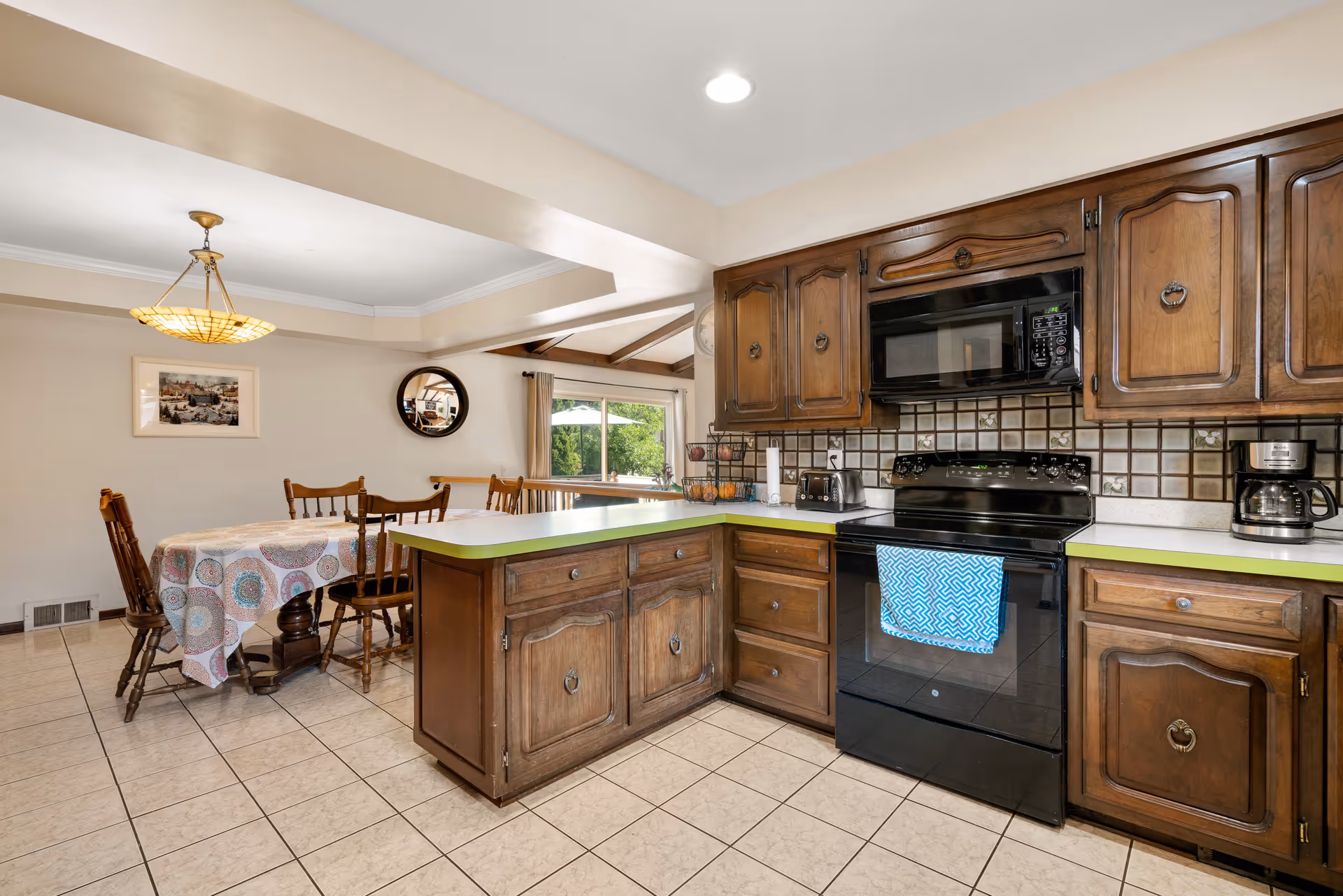A kitchen and dining area featuring wooden cabinets, a black stove with a blue patterned towel hanging on the handle, a microwave above the stove, a coffee maker, toaster, and a fruit basket on the counter. The dining area has a table covered with a patterned tablecloth and wooden chairs, with a window and a round mirror on the wall.