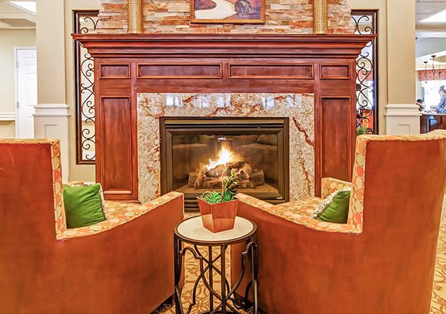 Two upholstered armchairs face a lit fireplace with a small round table and potted plant between them in a cozy lounge.