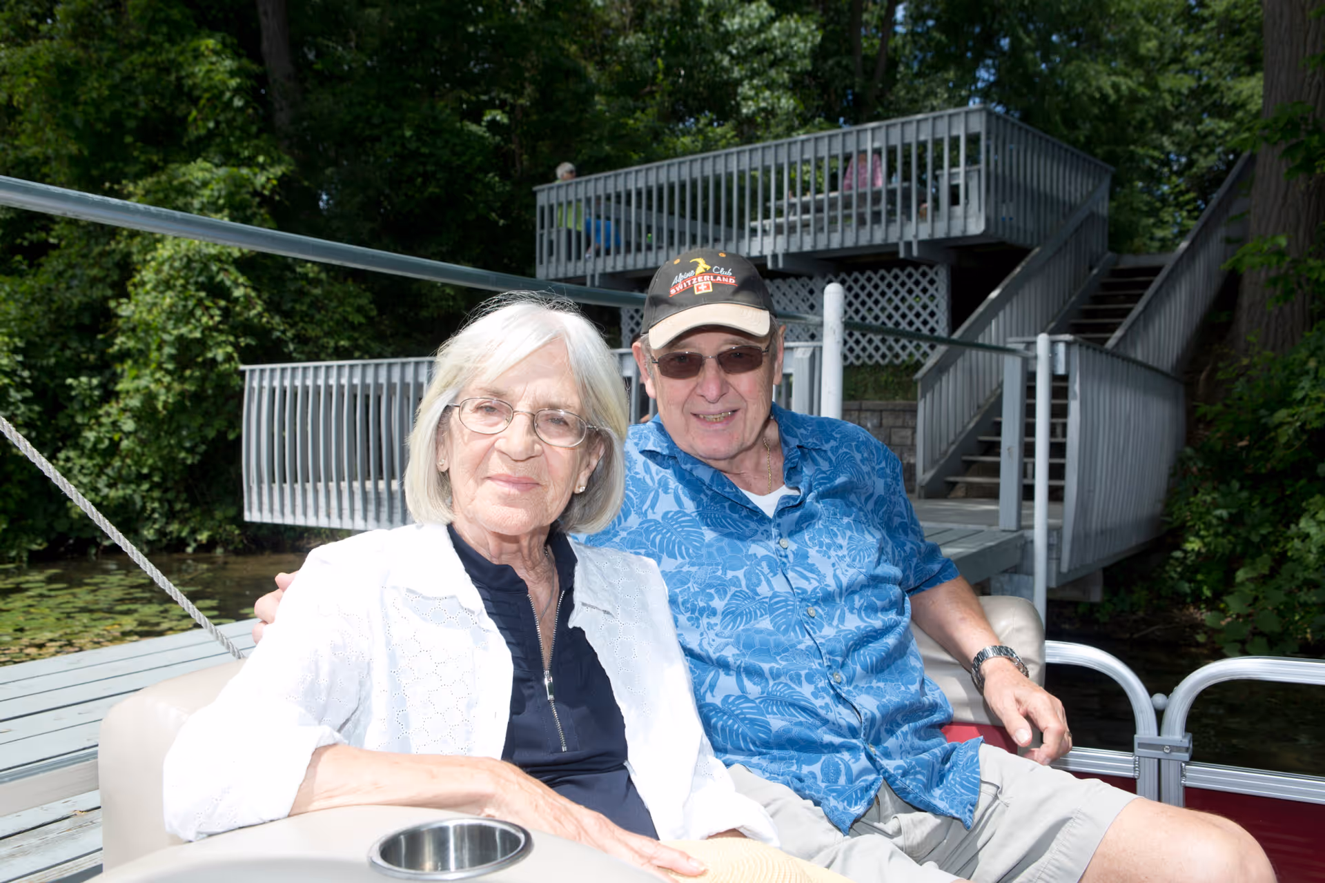 An elderly couple sitting together on a boat docked near a wooden deck surrounded by lush green trees. The woman has white hair and glasses, wearing a white jacket over a dark top. The man is wearing a blue patterned shirt, sunglasses, and a black cap. They are smiling and enjoying a sunny day outdoors.