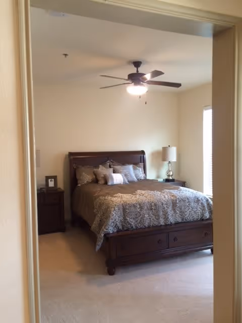 View through a doorway into a furnished bedroom with a wooden bed, patterned bedding, nightstands, a lamp, and a ceiling fan.
