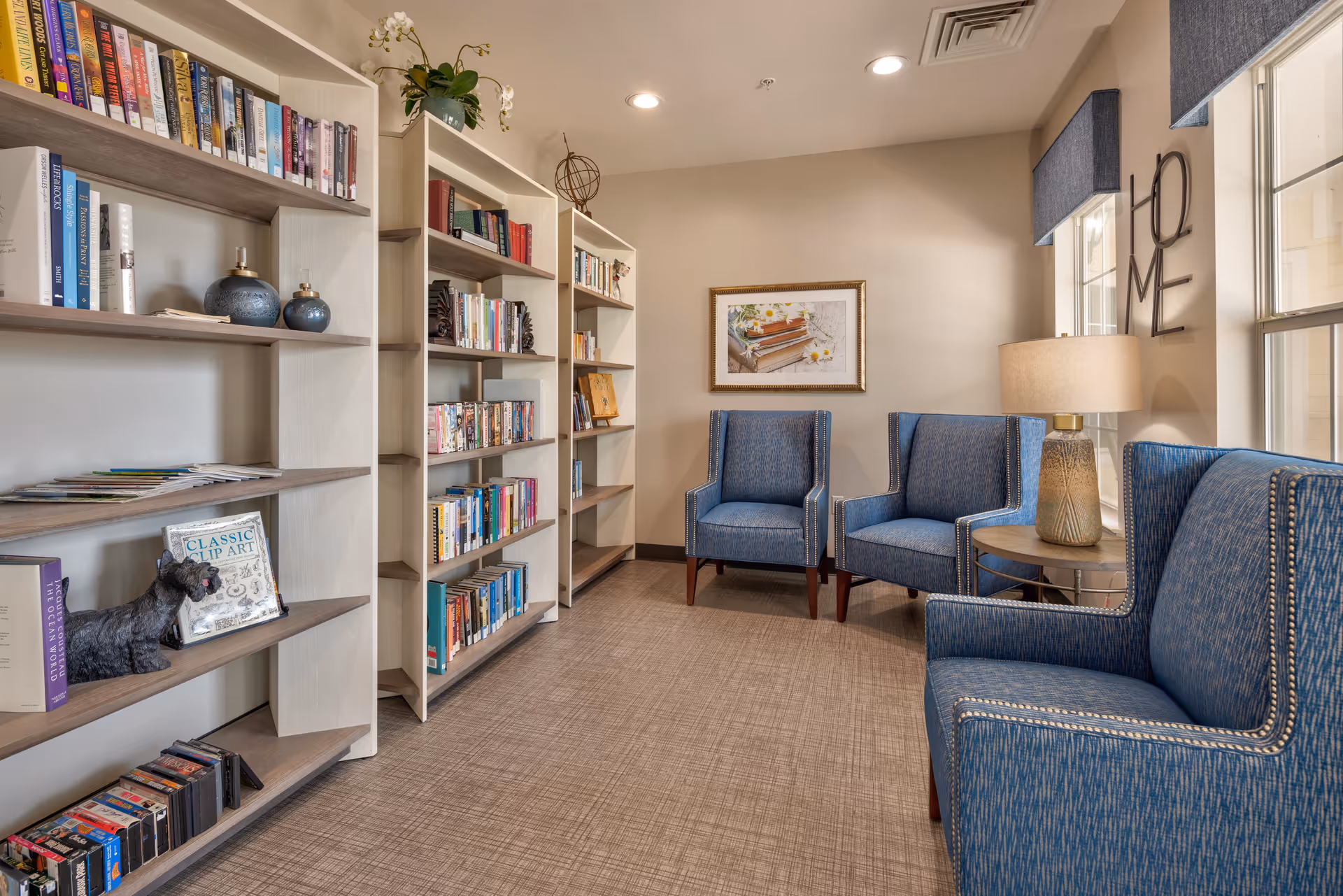 A cozy reading area in a senior living facility featuring multiple bookshelves filled with books and decorative items. There are three blue upholstered armchairs arranged around a small round table with a lamp. The walls are beige, with a framed picture and a decorative wall piece spelling 'HOME'. Two windows with blue valances allow natural light into the room.