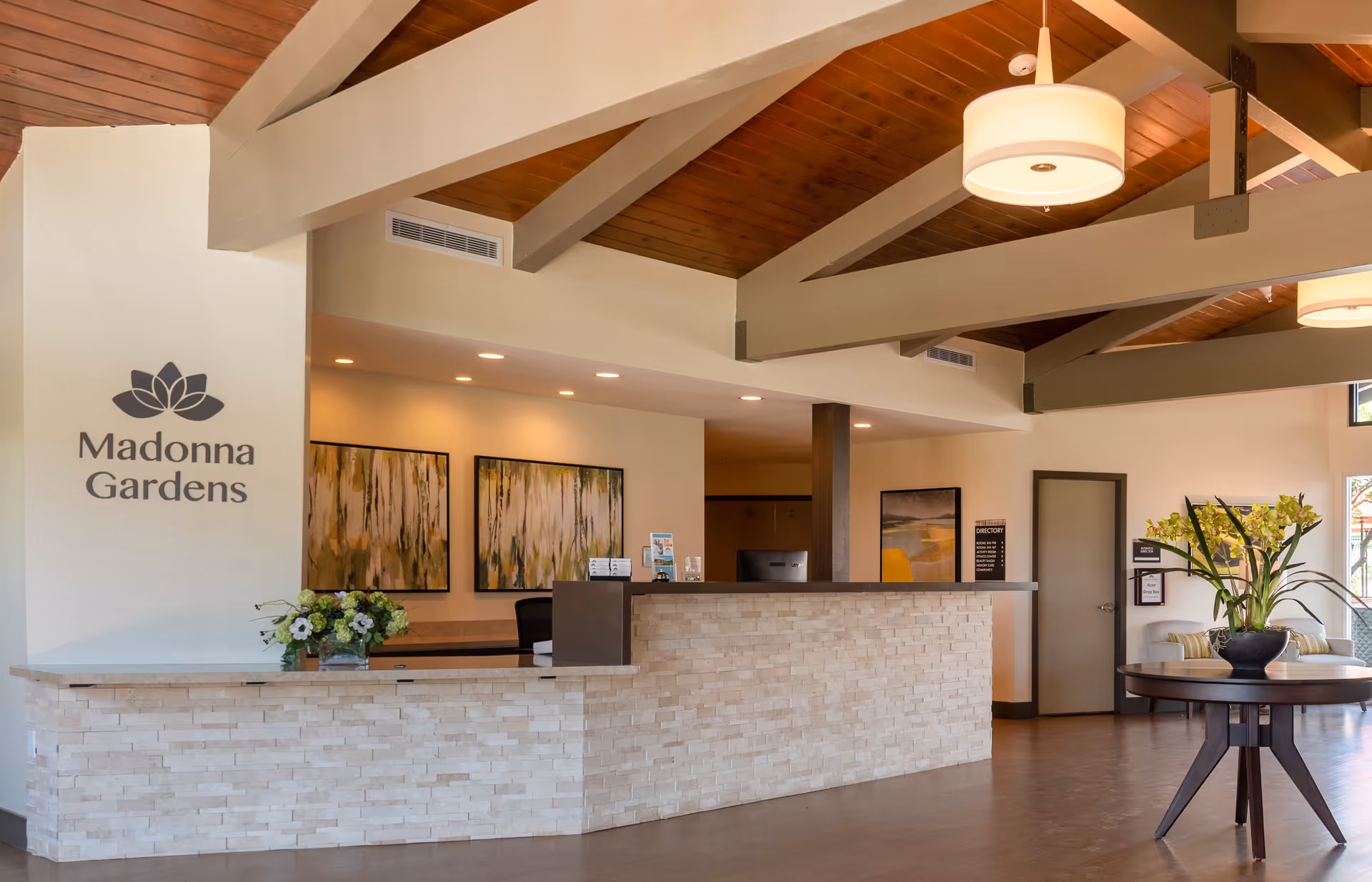 Reception area of Madonna Gardens Assisted Living and Memory Care featuring a modern stone front desk, two abstract paintings on the wall behind the desk, a vase with flowers on the desk, a round table with a large floral arrangement, wooden ceiling with exposed beams, and warm lighting.