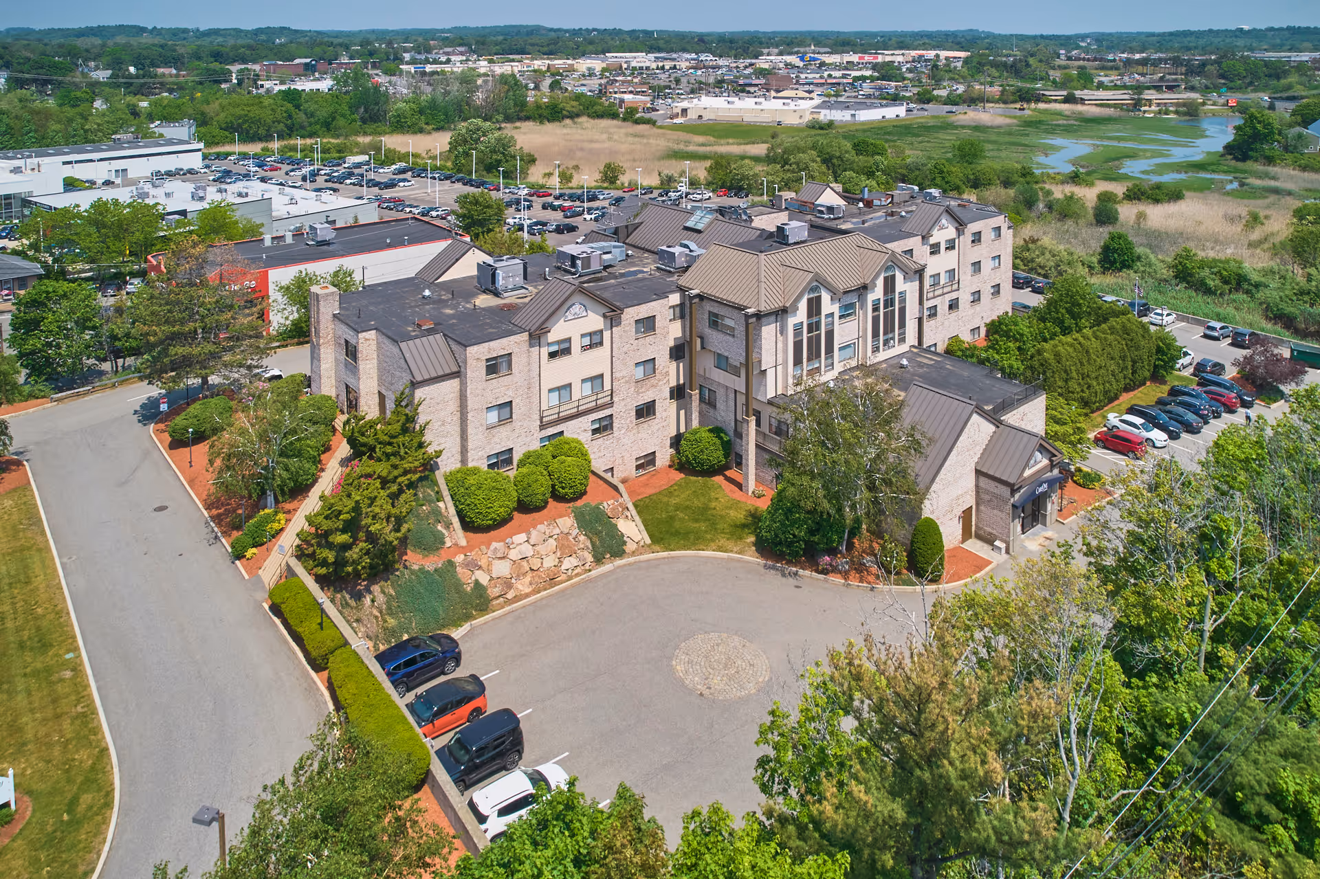 Aerial view of a multi-story senior living facility building surrounded by landscaped greenery, parking lots with cars, and nearby commercial buildings and wetlands in the background.