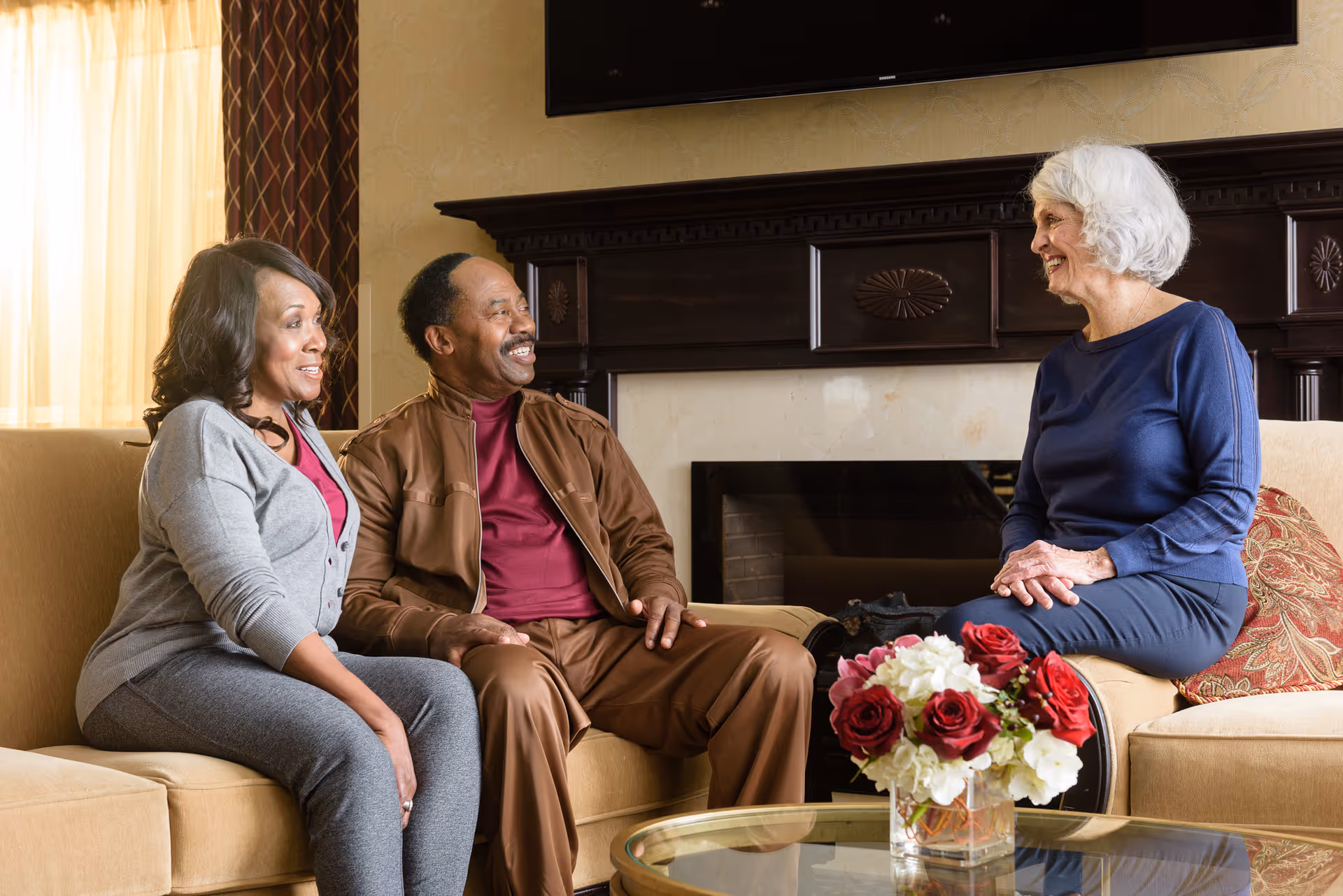 Three elderly people sitting and smiling in a living room. Two are seated on a beige couch on the left, and one is seated on a beige armchair on the right. There is a glass coffee table with a vase of red and white flowers in front of them, and a dark wooden fireplace mantle behind them.